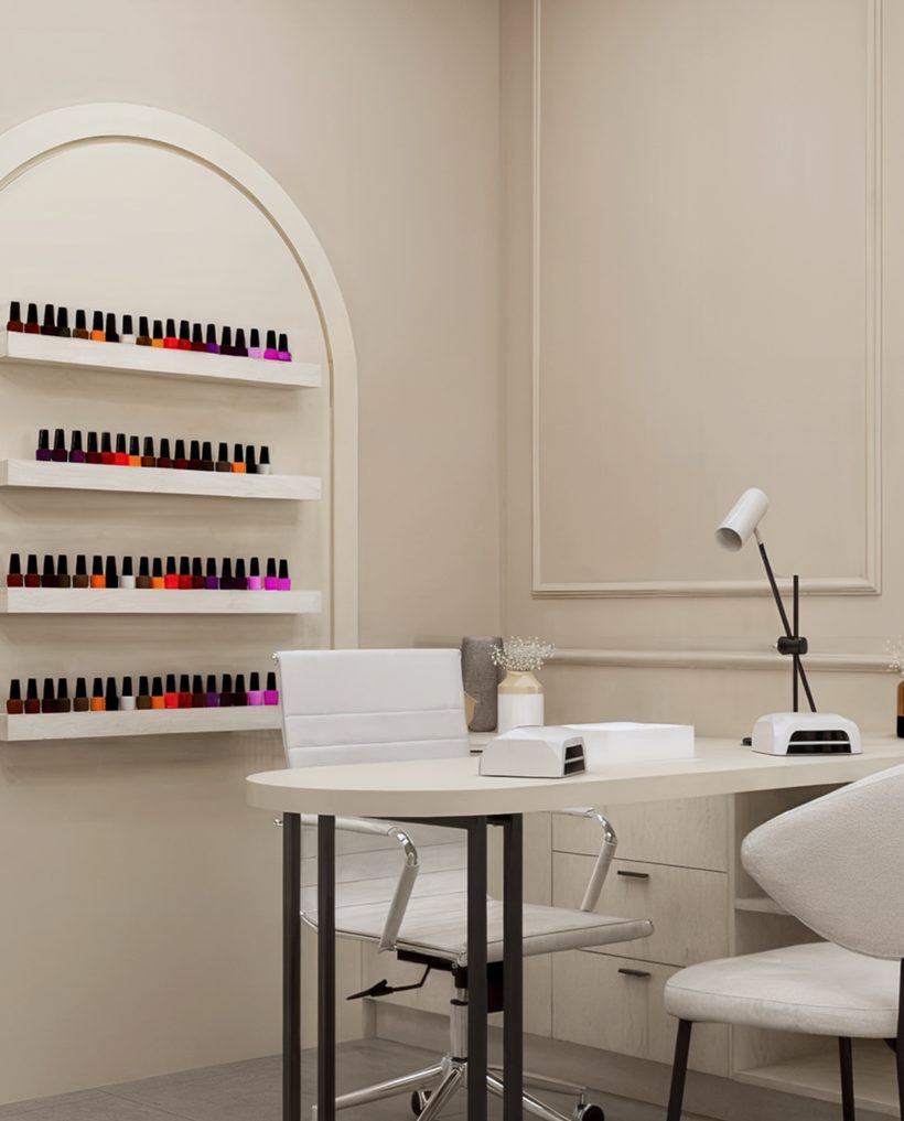 Mesa de manicure branca com luminária e cabine de LED, cadeiras claras e esmaltes em prateleiras num nicho em arco.